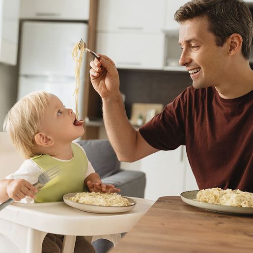 Vater füttert lachendes Kleinkind mit Löffel beim Essen am Tisch