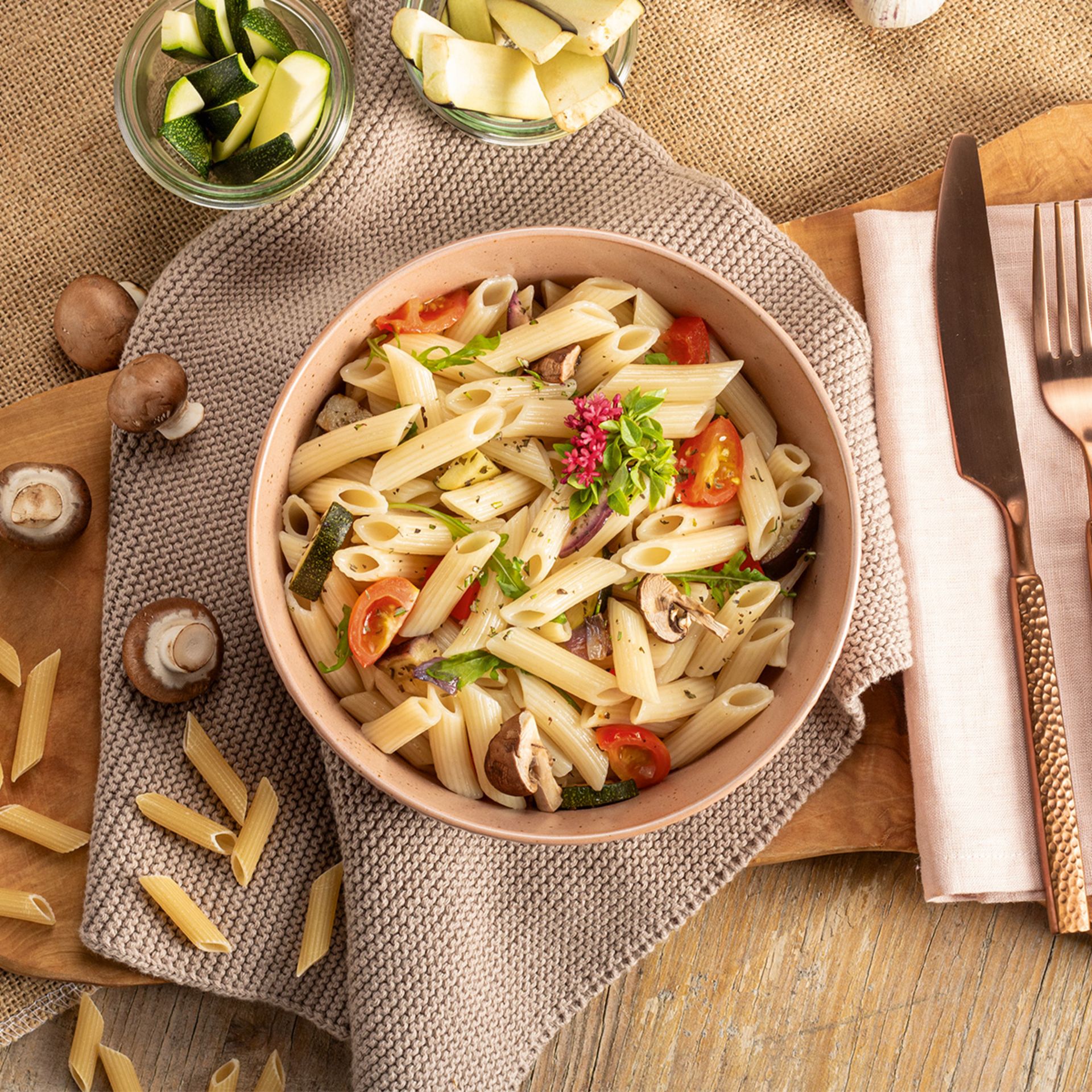 Nudelsalat mit Penne, Tomaten, Pilzen und frischen Kräutern in einer Schüssel auf einem grauen Tuch
