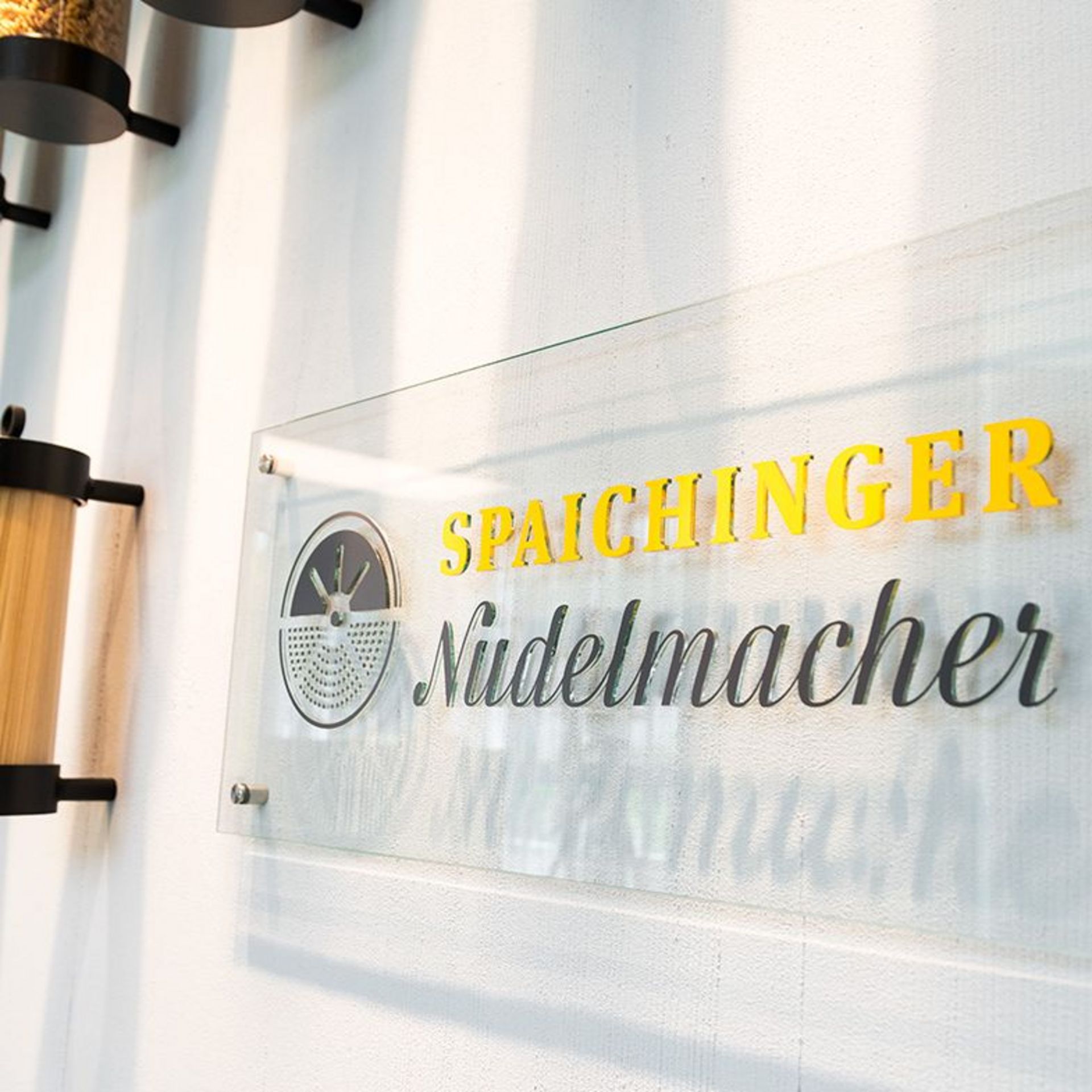 Transparentes Schild mit gelbem Schriftzug 'Spaichinger' und schwarzem Schriftzug 'Nudelmacher' an einer weißen Wand
